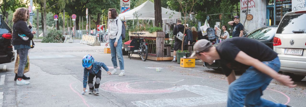 Ein Kind und ein Erwachsener malen mit bunter Kreide Zahlen auf eine Fahrbahn.