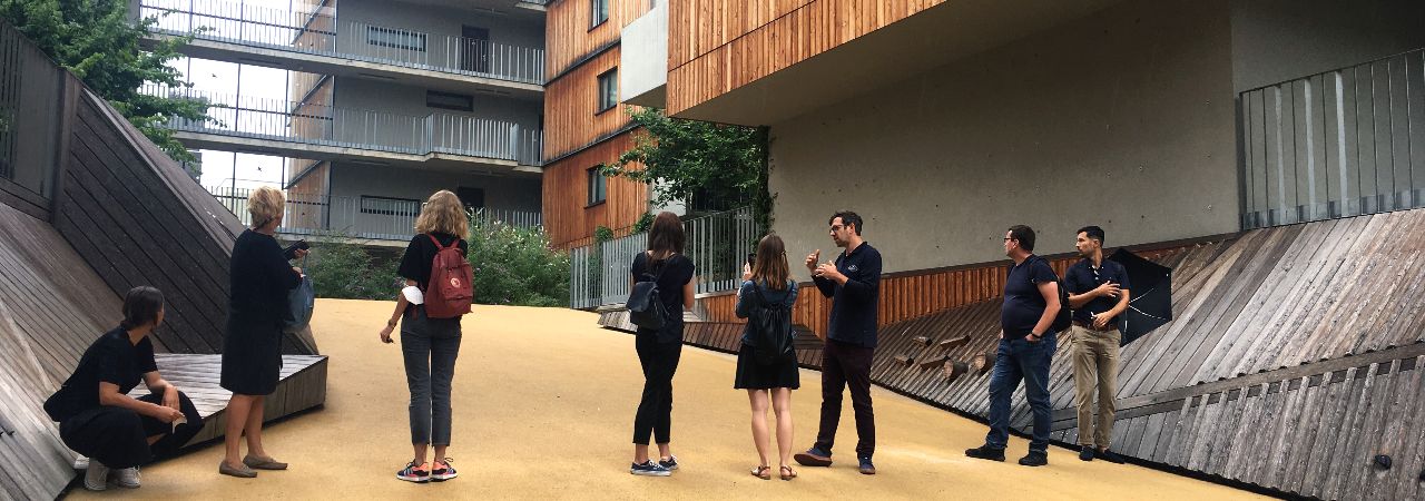 Mehrere Personen stehen vor einem Wohngebäude aus Holz, im Hintergrund ein Betongebäude